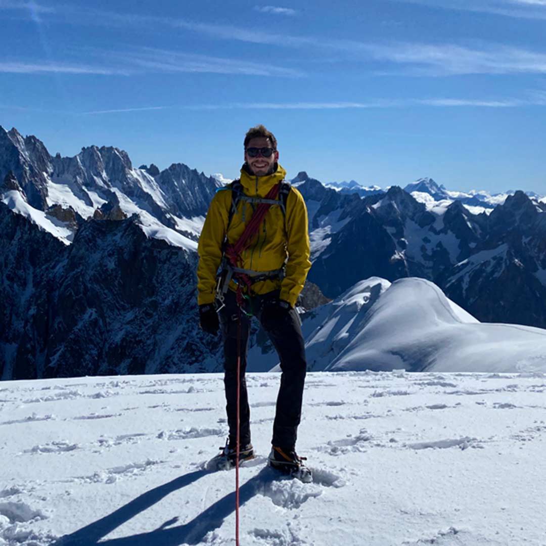 A man stood infront of a snowy mountain vista wearing climbing equipment.