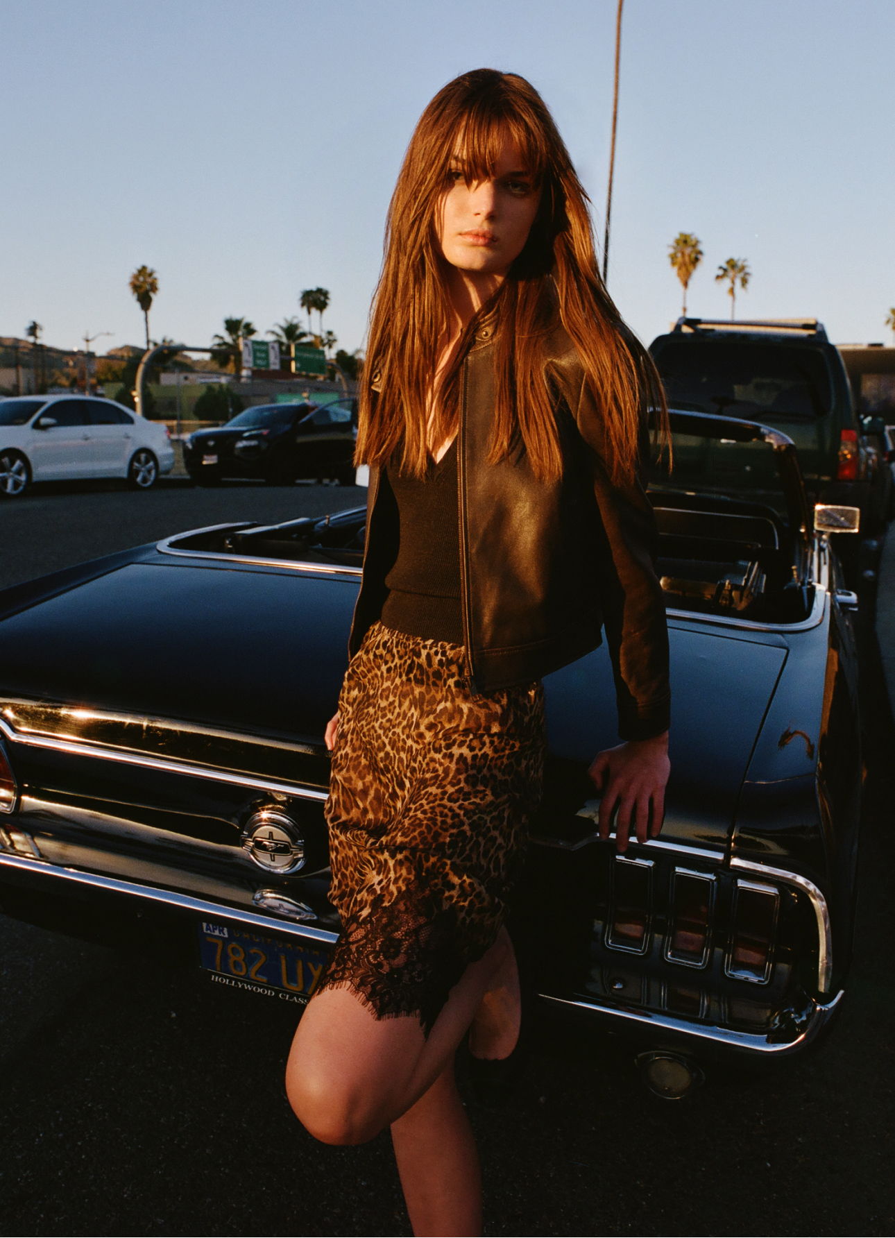 Model in a leather jacket poses by a vintage car at sunset.