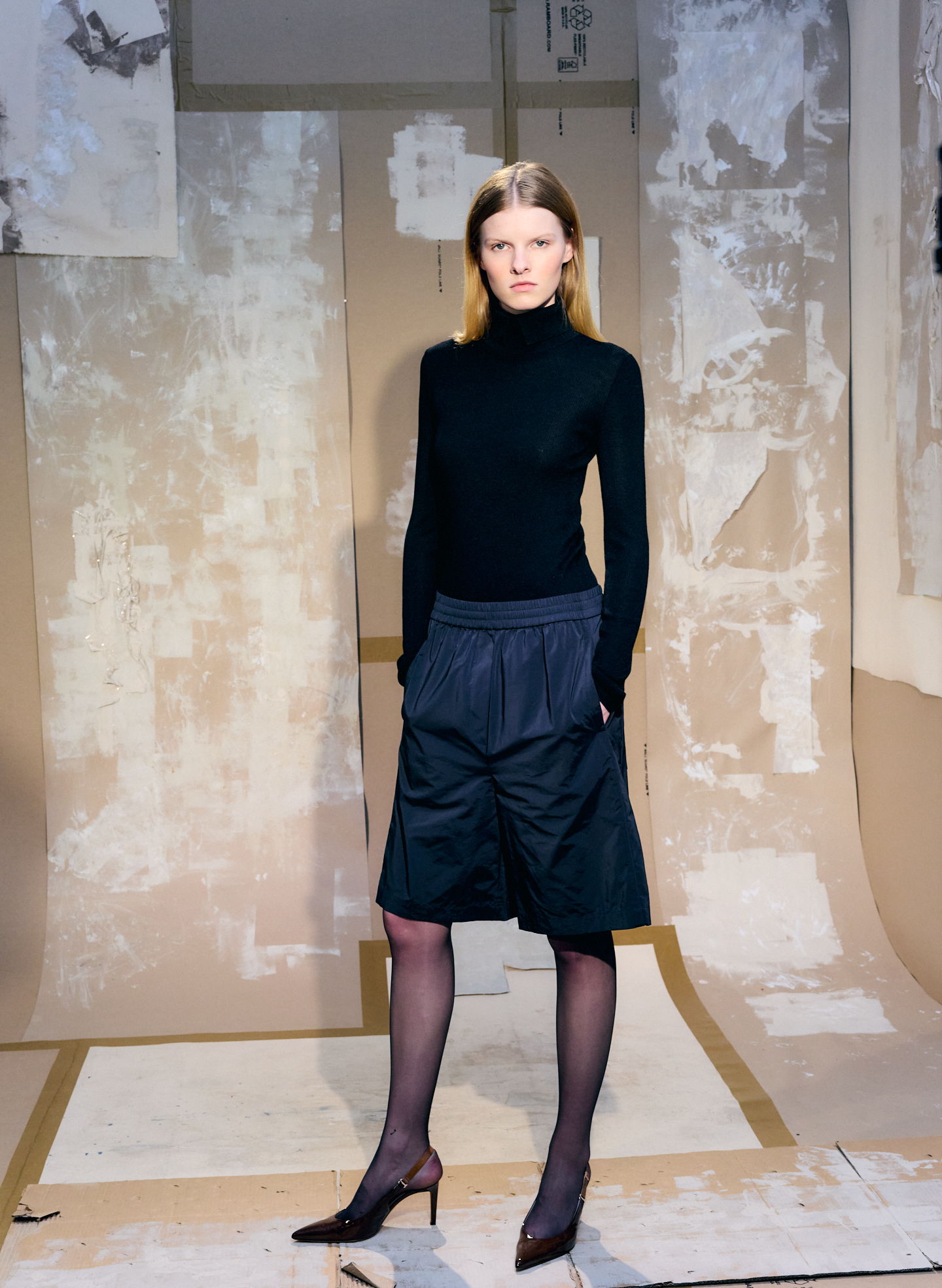 Model in black turtleneck and skirt poses against a textured background.