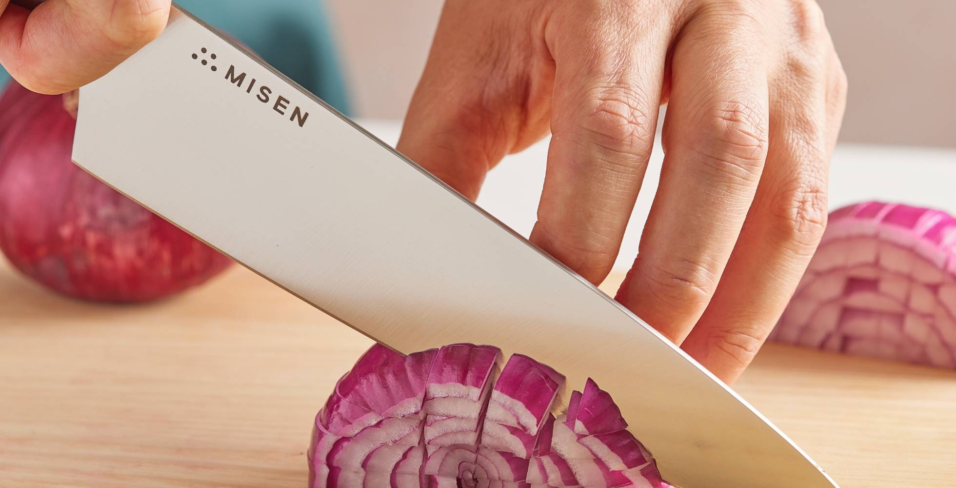 Hands use a blue Misen Chef’s Knife to dice red onions on a light wood cutting board.