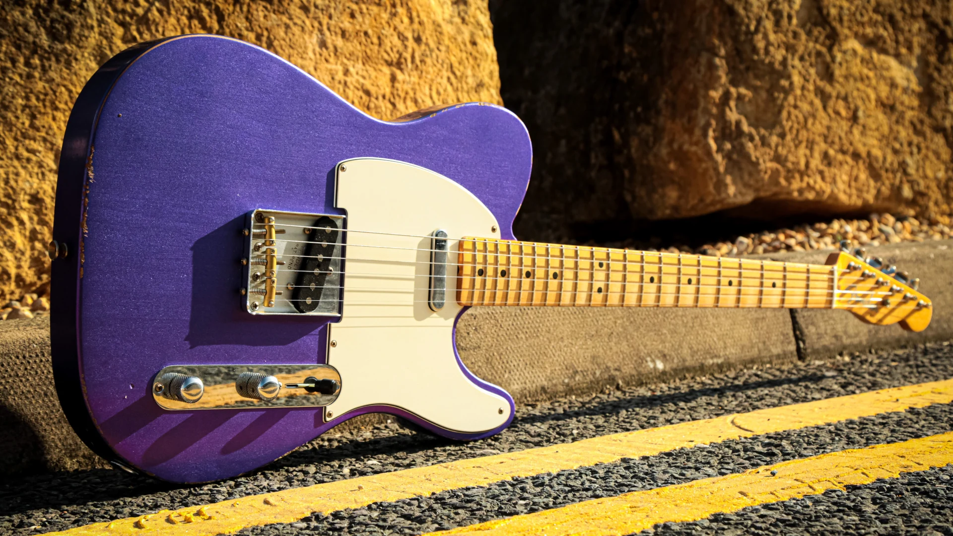 Purple Fender Telecaster electric guitar resting on a road beside a rocky surface.