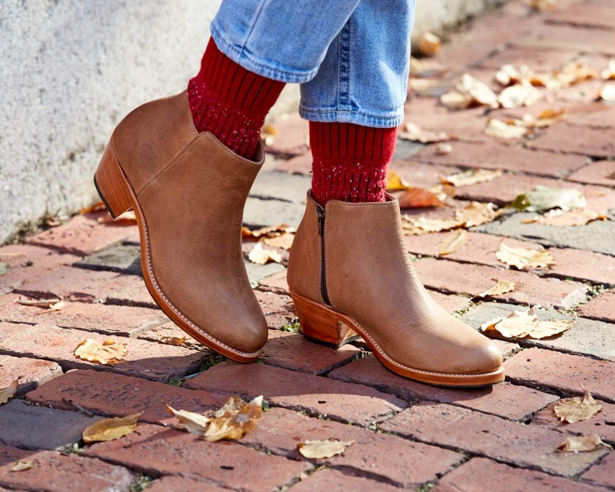 Close-up view of a person wearing the handcrafted Adelante Granada leather ankle boots in desert brown, walking on a brick pathway scattered with autumn leaves.