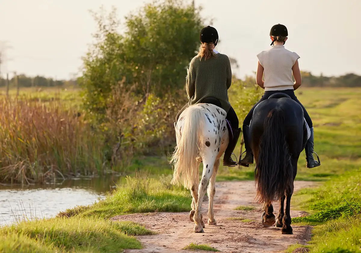 Two riders on horseback walking along a path beside a river.