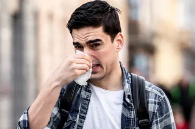 Man in wit T-shirt en blauw overhemd snuit zijn neus in een zakdoek. Dit is een typisch symptoom van allergie. Veel niezen en snuiten door een loopneus of een verstopte neus.