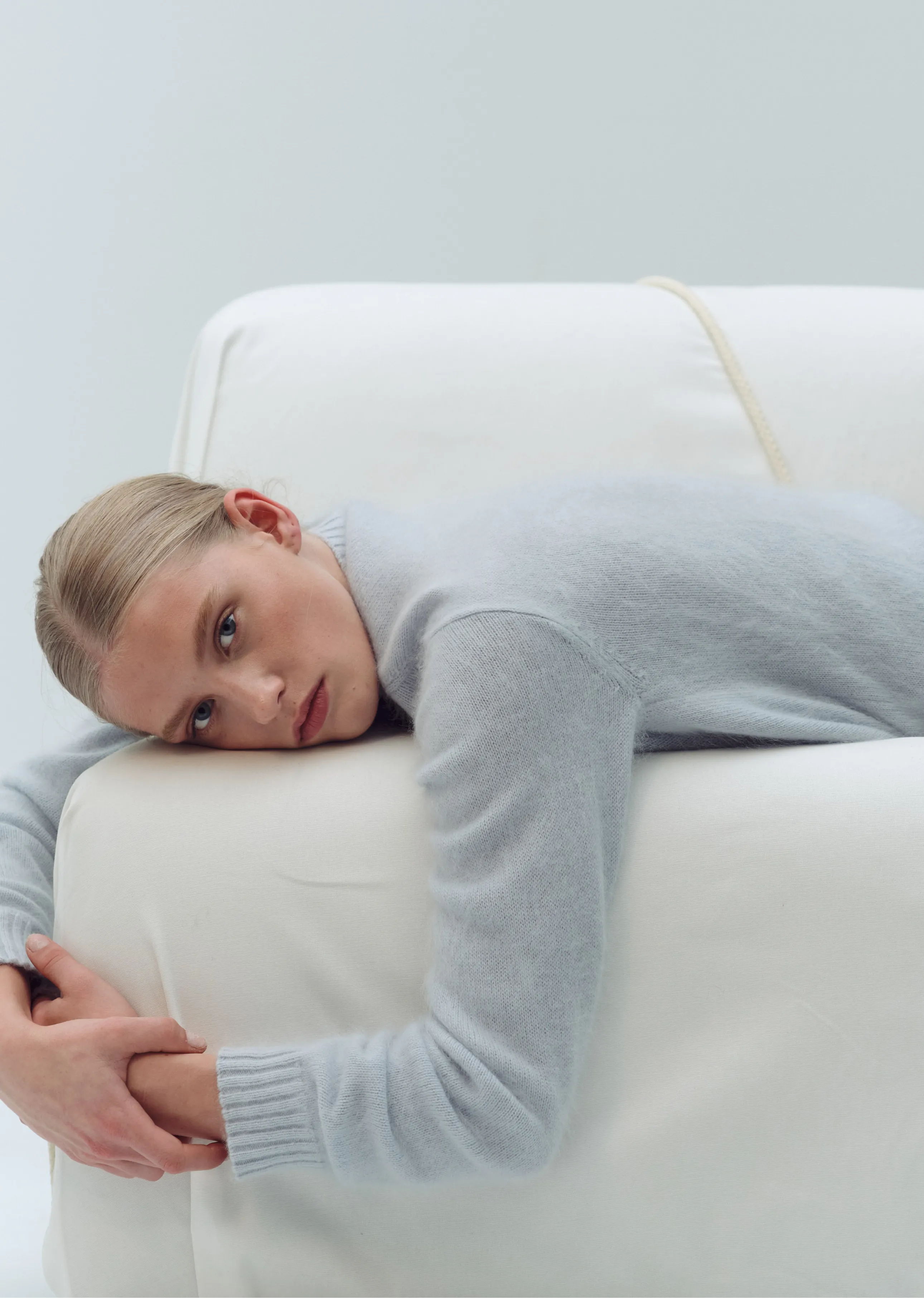 Person laying on a white couch, looking contemplative in a light sweater.