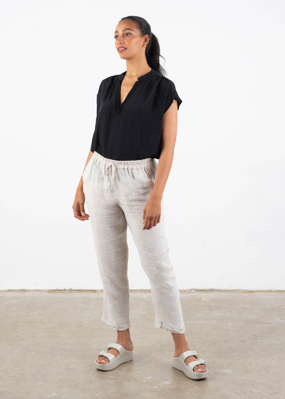 A model wearing a pear of taupe coloured tapered linen trousers with a black short sleeved top and off white chunky platform slides