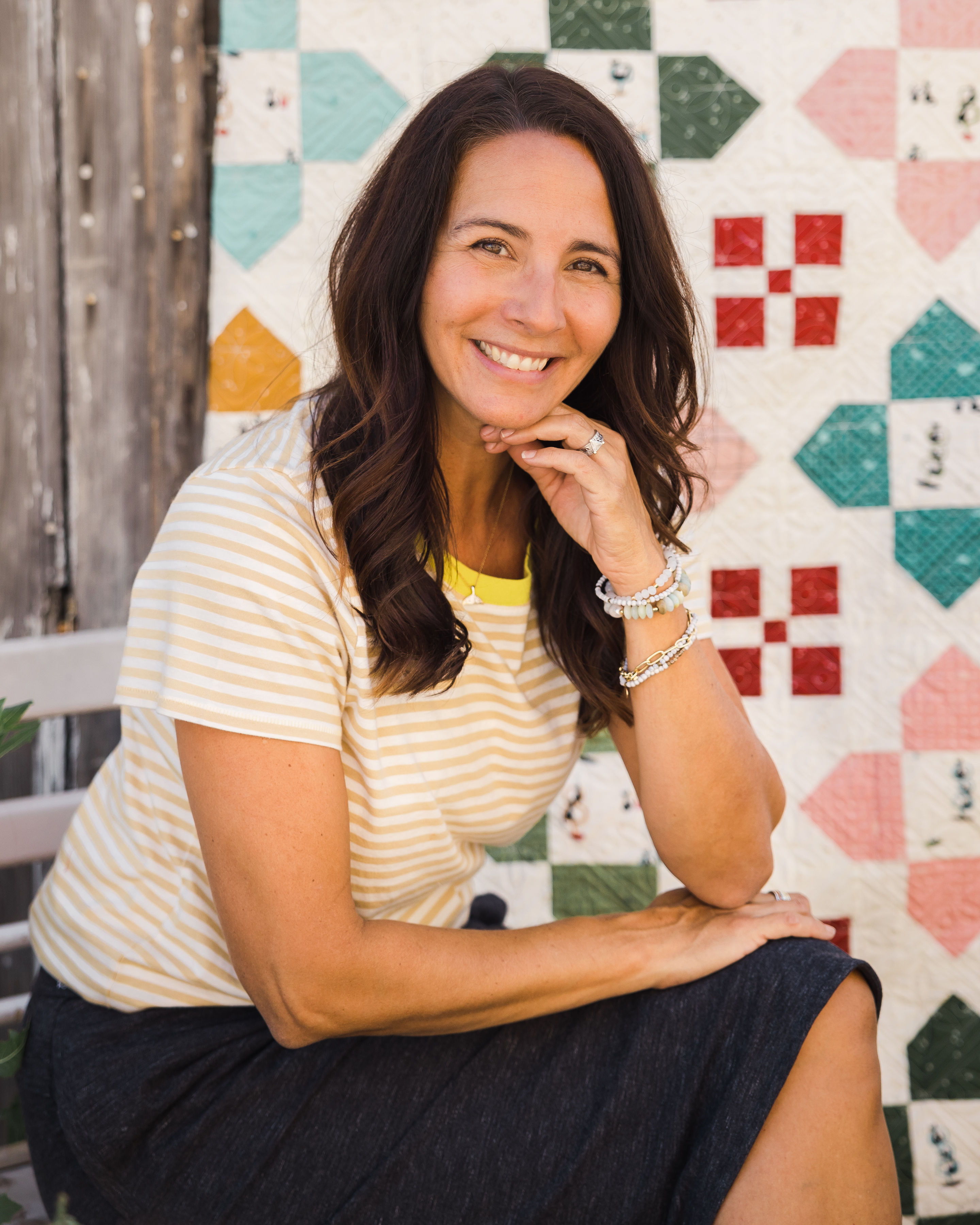 Smiling woman in striped shirt sitting against a colorful quilt backdrop.