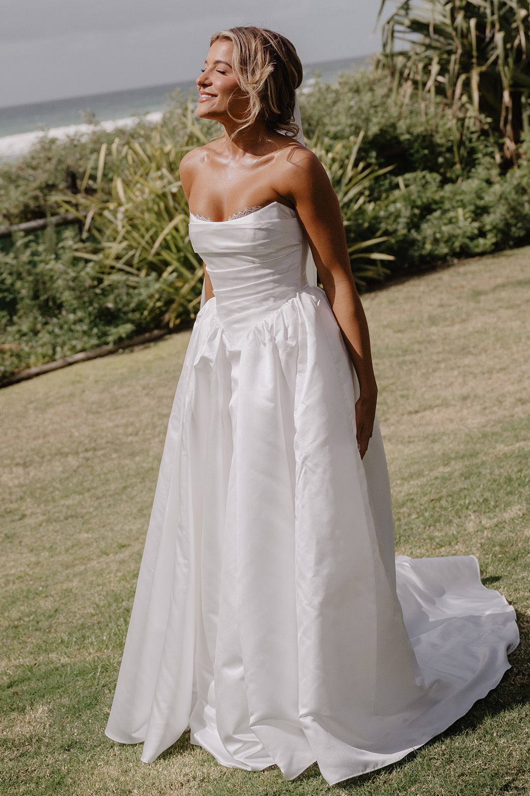 Woman in a white wedding dress standing on grass near the beach.