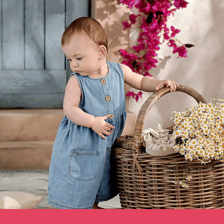 Baby in a denim dress beside a wicker basket with flowers.