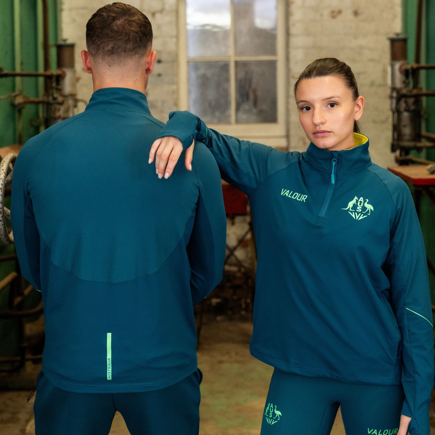 Two athletes in matching teal jackets, one facing forward, one facing back.