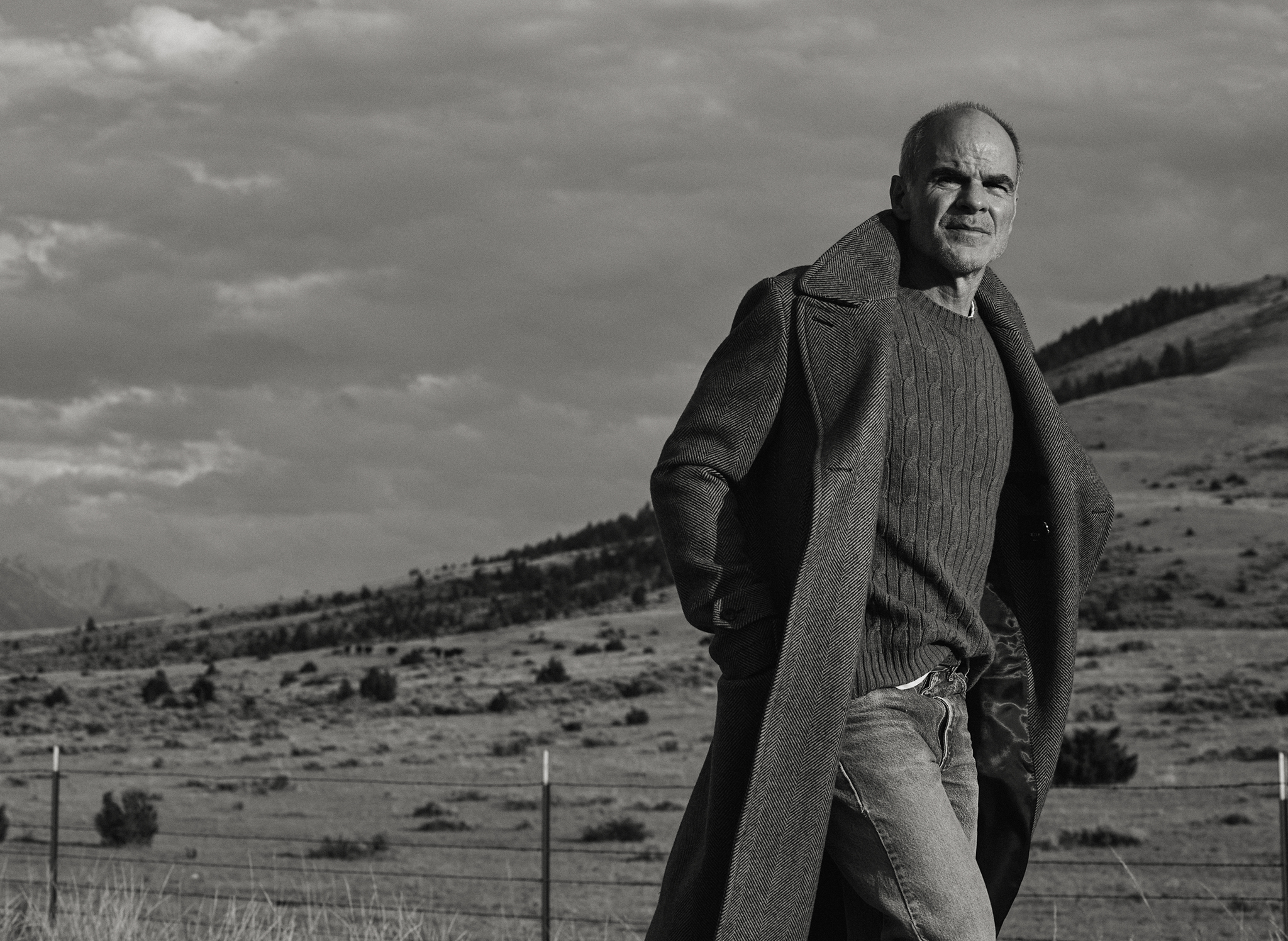 Michael Kelly in a black and white photograph wearing the Barrow Wool Herringbone Double Breasted Topcoat in a Montana Field with a barbed wire fence and mountains in the background
