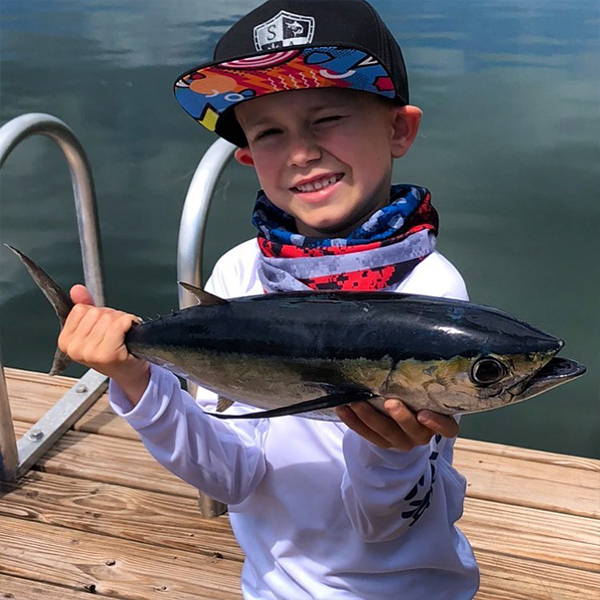 Bosun Lindsey holding up a fish smiling at the camera while wearing a kid's performance shirt, a face shield around his neck and an SA Company hat.
