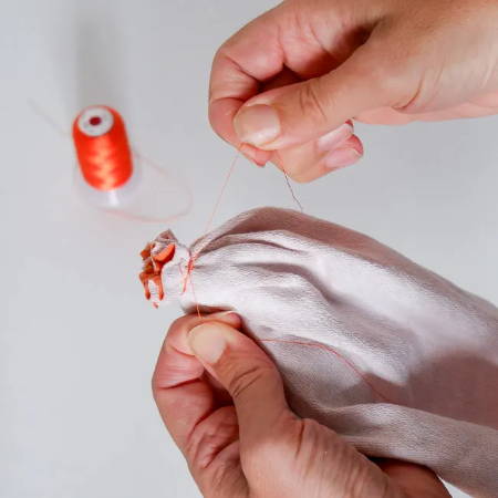 Hands pulling orange thread to gather the fabric of a fabric tube and tying a knot
