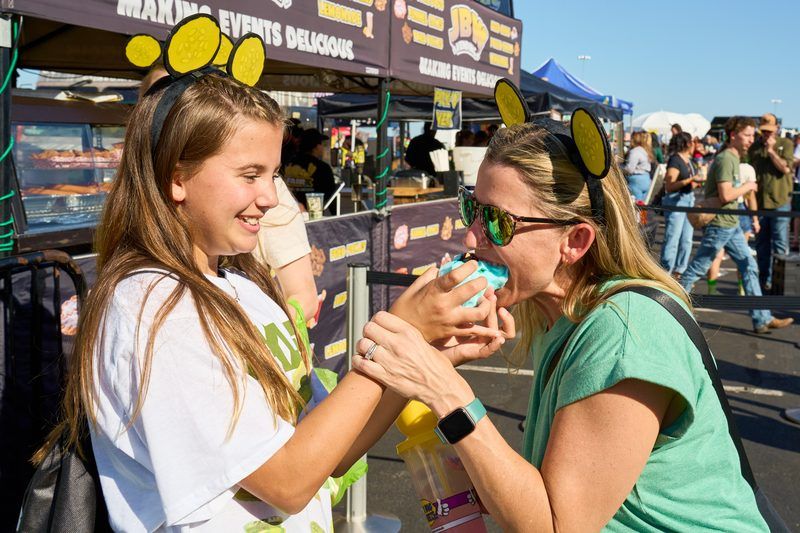 Mother and daughter in pickle ear headbands sharing cotton candy in front of the fried pickle and funnel cake food vendors, an all-ages moment at The Big Dill® Philadelphia pickle party at Stateside Live! on National Pickle Day