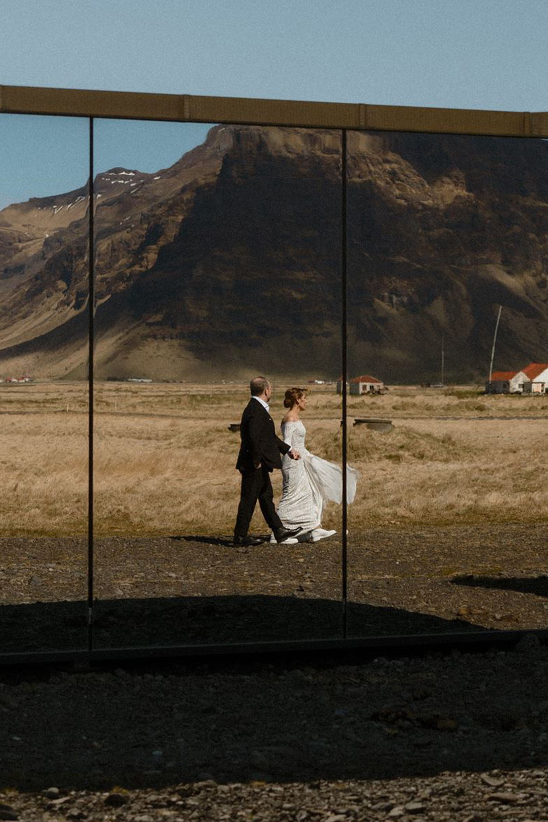 A couple walks together, reflected in a large mirror with mountains in the background.