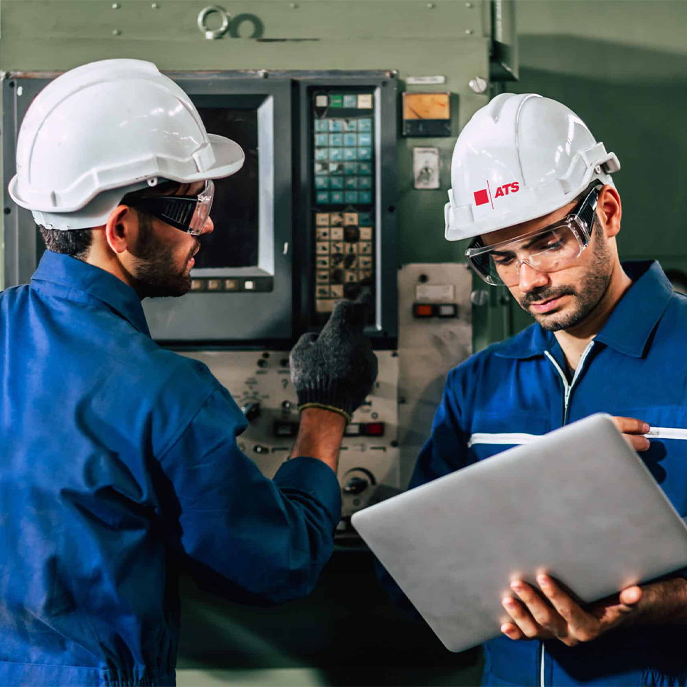 Two workers in hard hats examining machinery and using a laptop.