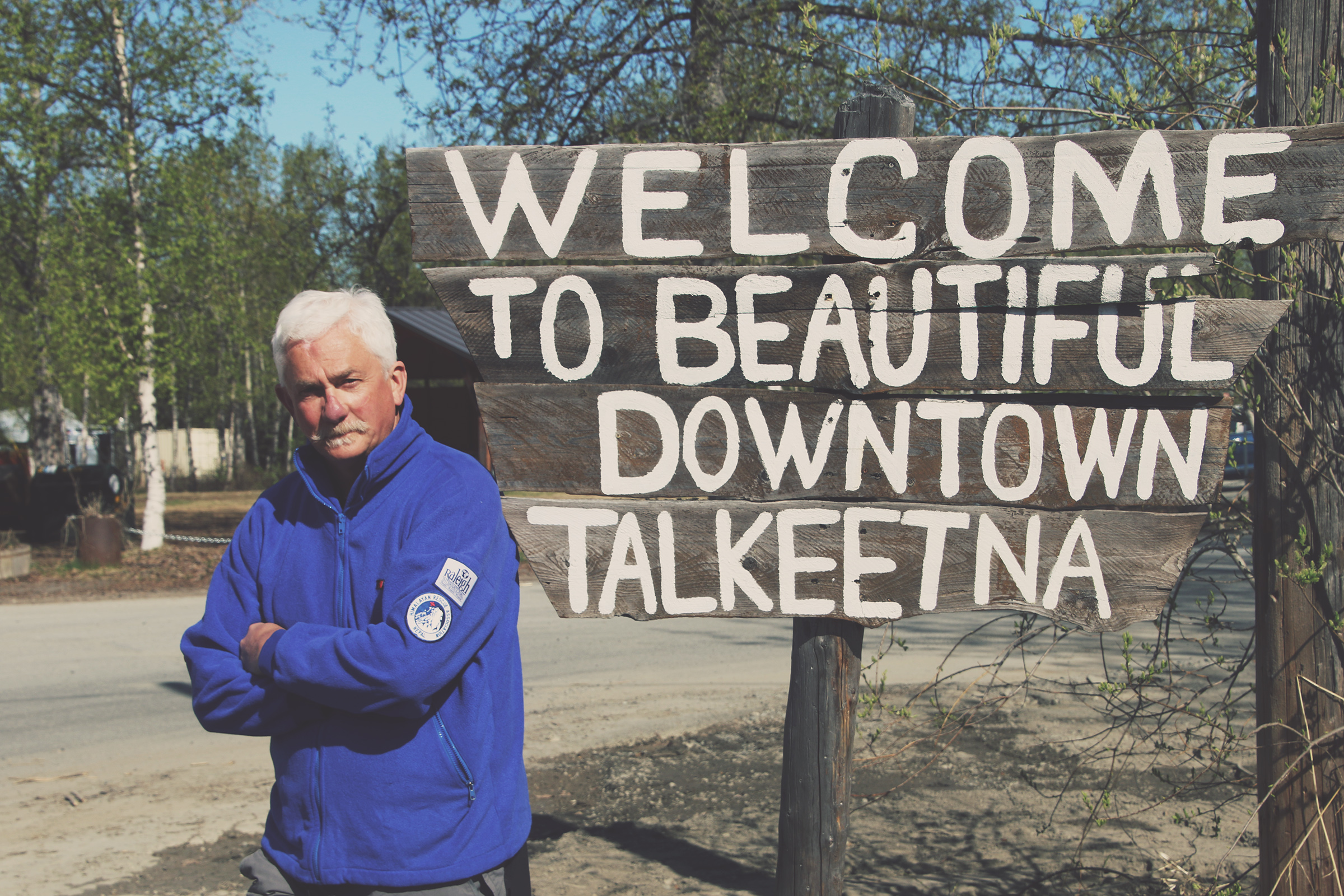 Man in blue jacket next to a sign welcoming to Talkeetna.