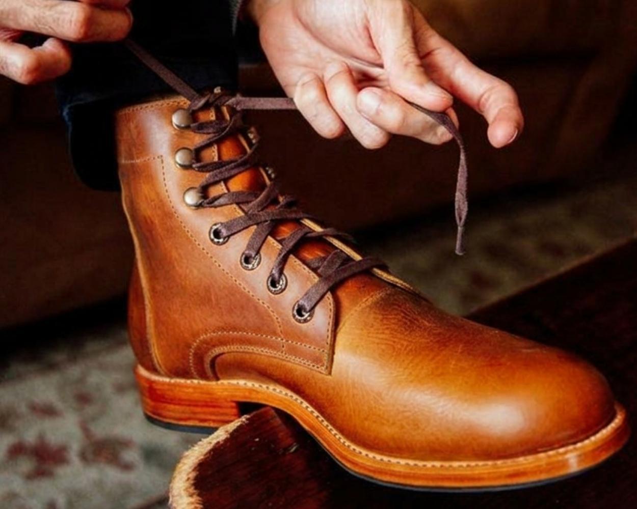 A close-up shot focusing on a single, high-top tan or caramel brown leather Adelante Havana lace-up boot. A person's hands are shown tying the dark brown laces near the top speed hooks. The boot features detailed Goodyear welt stitching along the sole edge and a stacked leather heel, resting on a dark wood surface or step with a warm, indoor setting blurred in the background.
