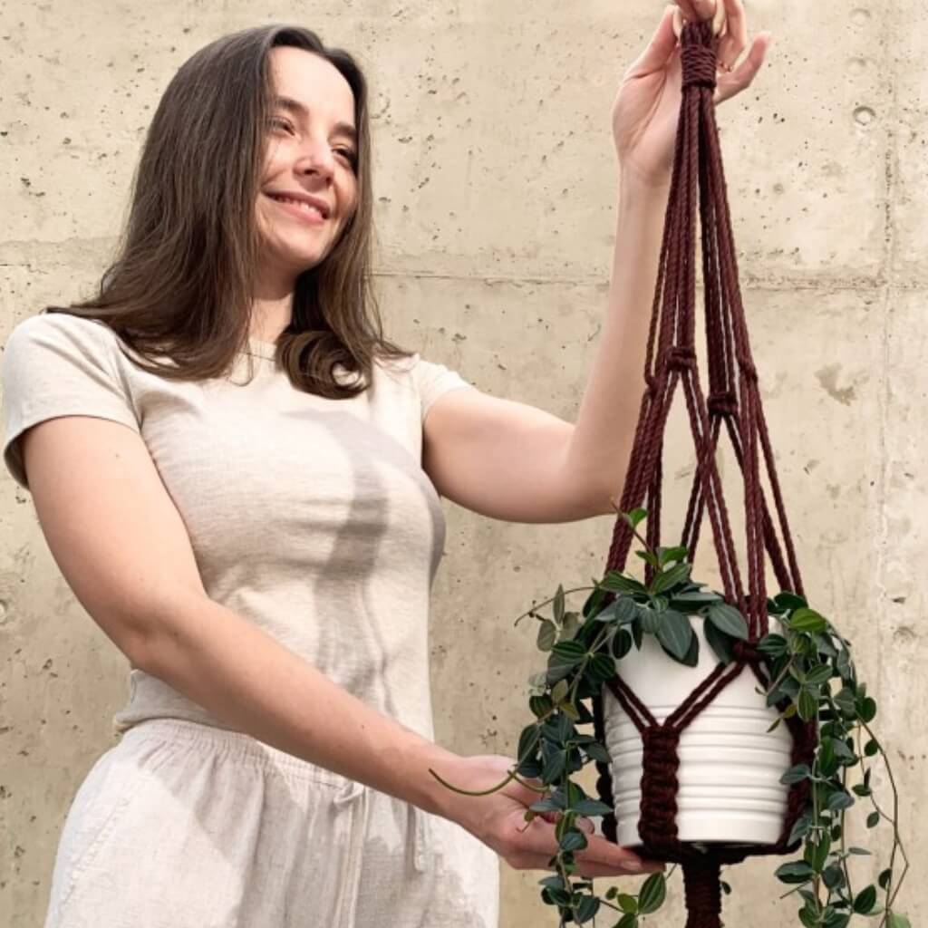 Eine Frau hält eine Makramee Blumenampel mit Grünpflanze vor einer strukturierten Wand.