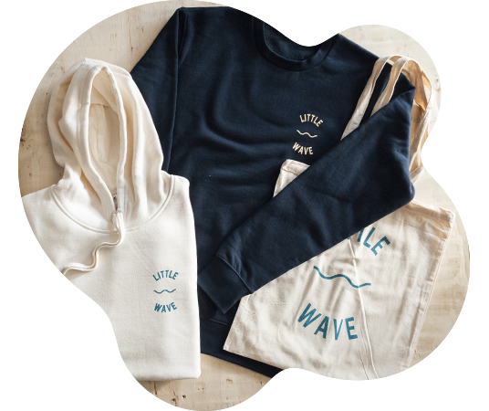 Flat lay of cream and navy Little Wave hoodies and tote bag, screen printed with the Little Wave logo and wave motif