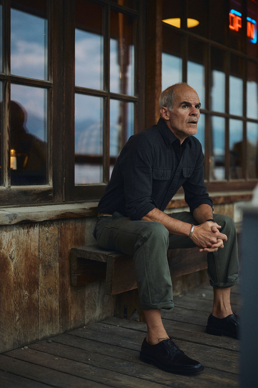 Michael Kelly sitting on a bench outside the Old Saloon at dusk wearing the Rivay Miles Brushed Flannel Shirt and Series II Garment Dyed Utility Pant in Vintage Olive