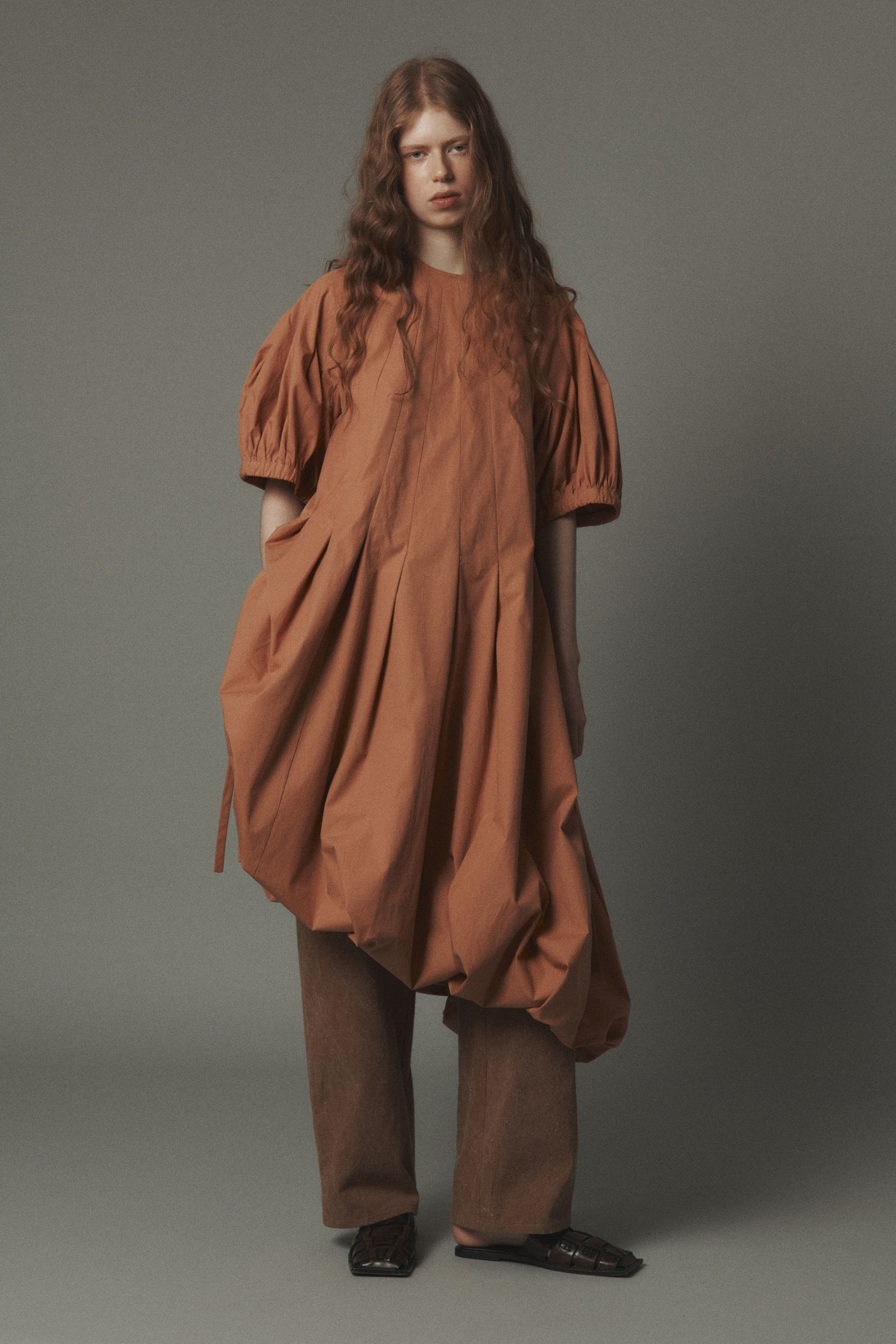 Model wearing oversized brown dress and loose pants against a gray background.