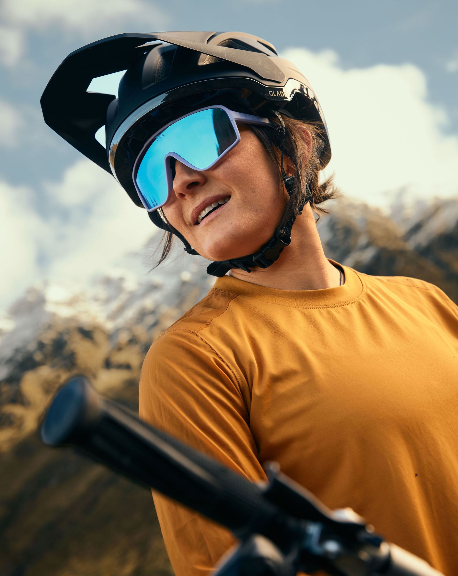 Person wearing a helmet and sunglasses, smiling outdoors with mountains in background.