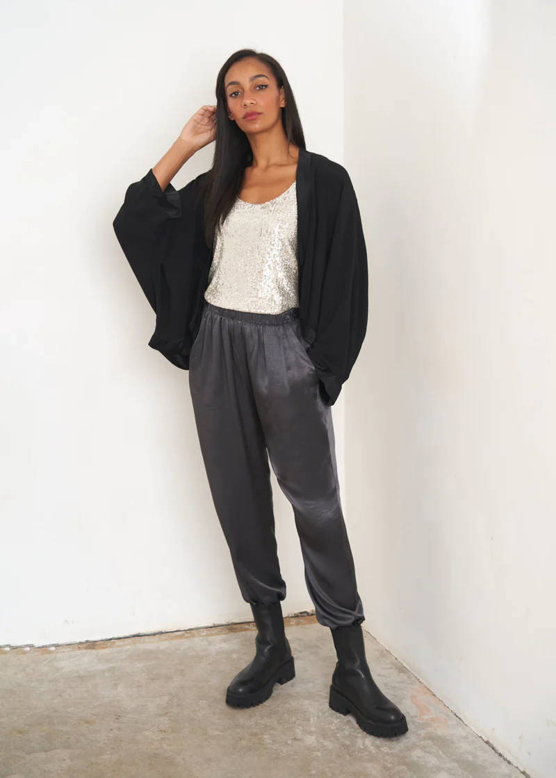 A model wearing a black kimono style jacket over a sequin silver top, dark grey satin trousers with black chunky chelsea boots