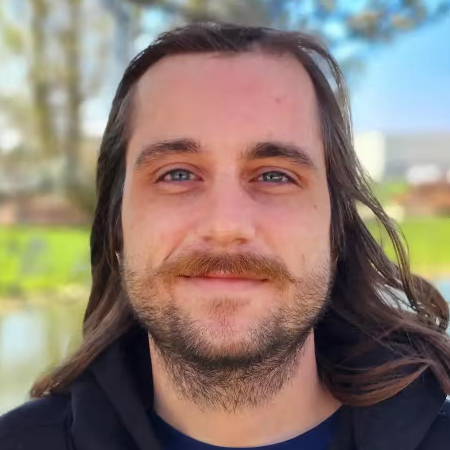 Smiling individual with long hair, wearing a black hoodie, near a body of water.