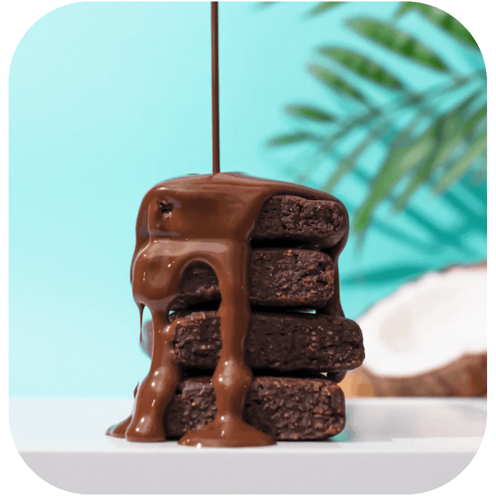 Stack of brownies with chocolate drizzling over them. Light blue background with palm fronds and coconut in background