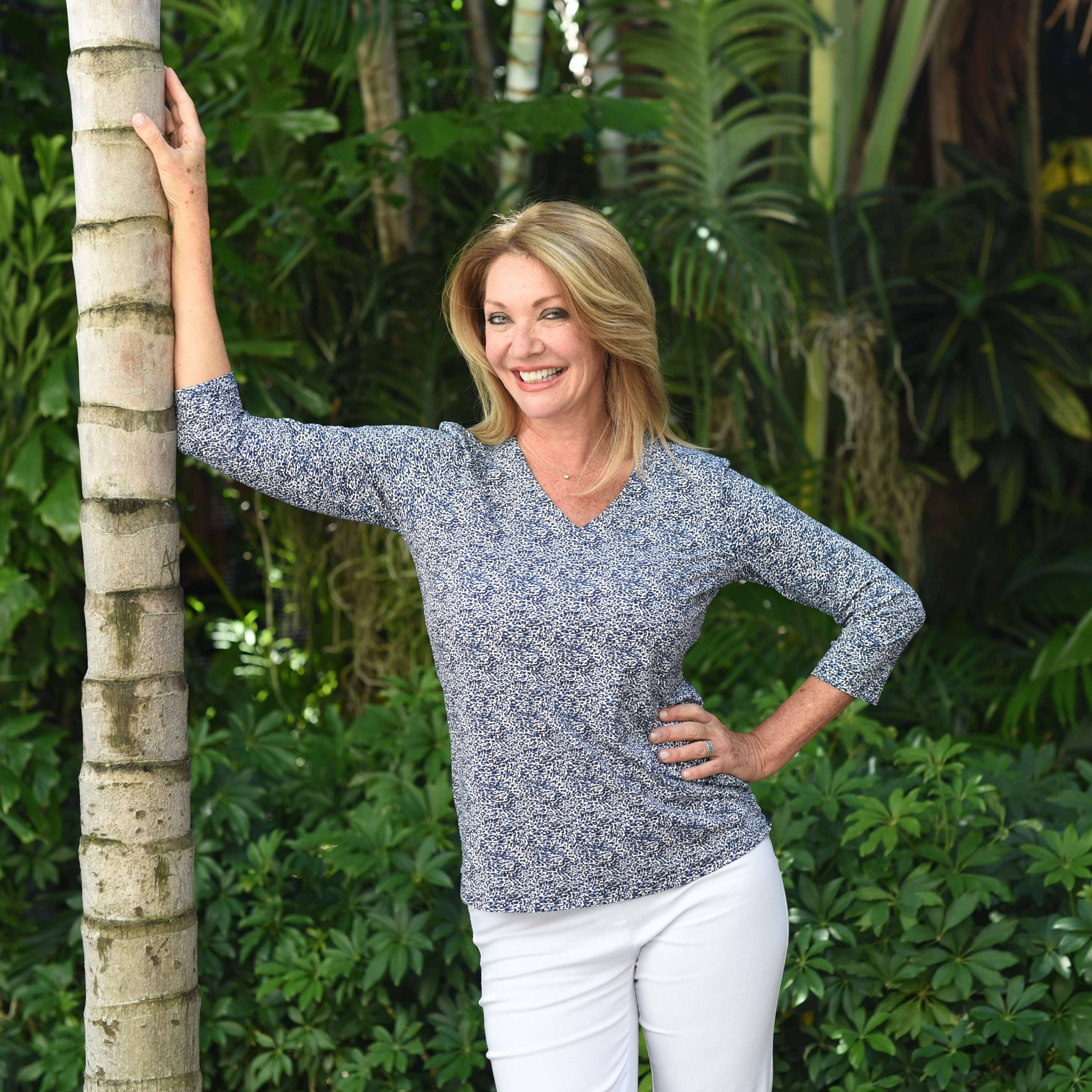A blonde smiling blonde woman leans against a palm tree. 
