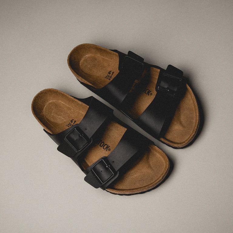 A pair of black sandals with two straps on a neutral background.