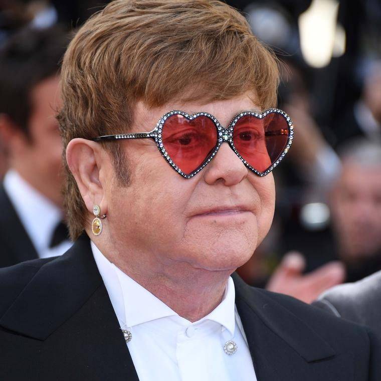 Sir Elton John porte des lunettes de soleil en forme de cœur avec des verres roses, une boucle d'oreille dorée avec une chemise blanche et une veste noire