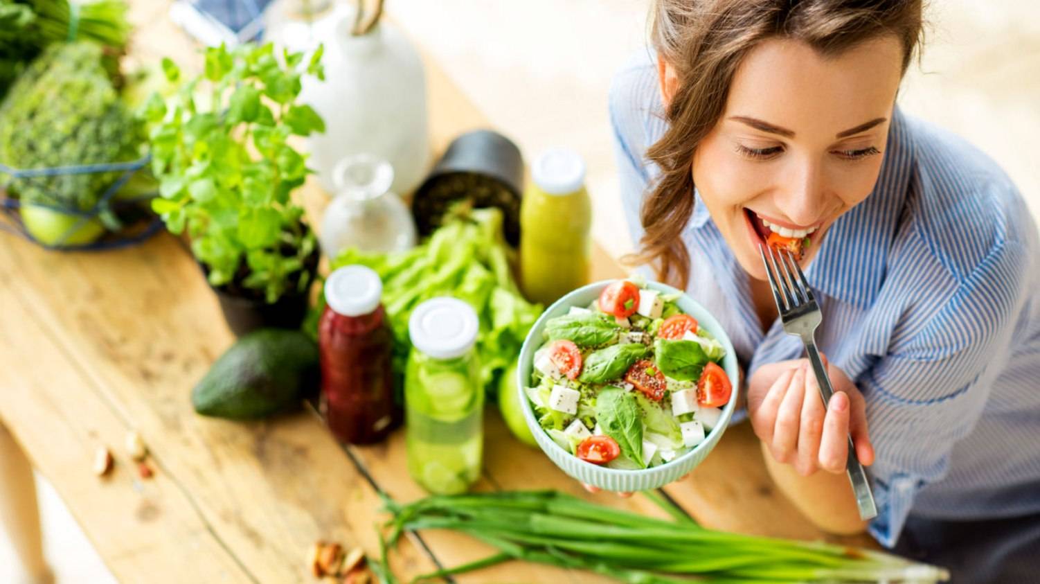 Featured | Young and happy woman eating healthy salad sitting on the table with green fresh ingredients indoors | How To Get Started On Vegan Keto Diet - The Ultimate Guide