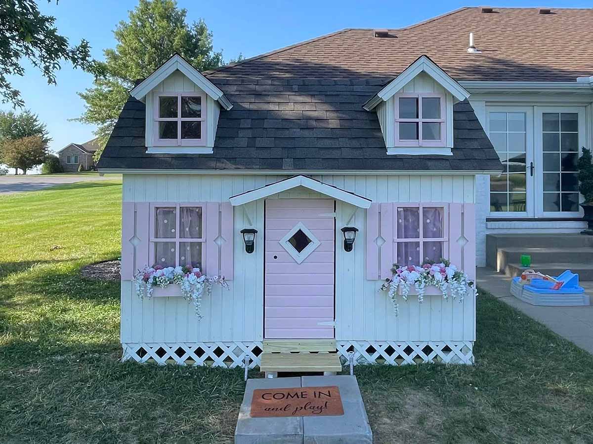 White and Pink Farmhouse style Playhouse with pink Flower boxes and window shutters in a backyard by WholeWoodPlayhouses