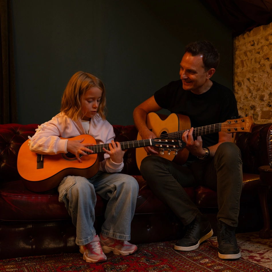 A girl and a man playing acoustic guitars on a sofa