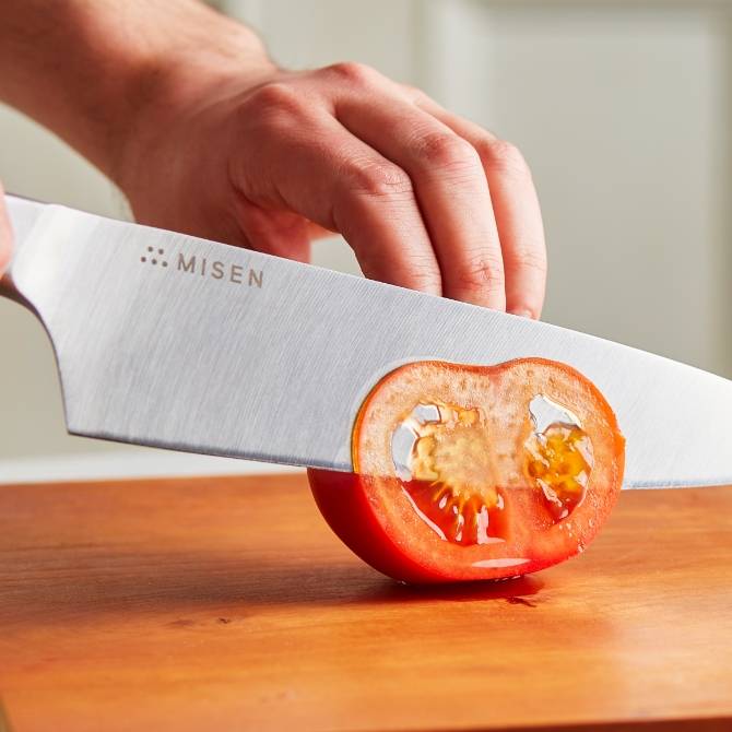 Hands use a blue Misen Chef’s Knife to cut a nearly transparent slice of tomato.