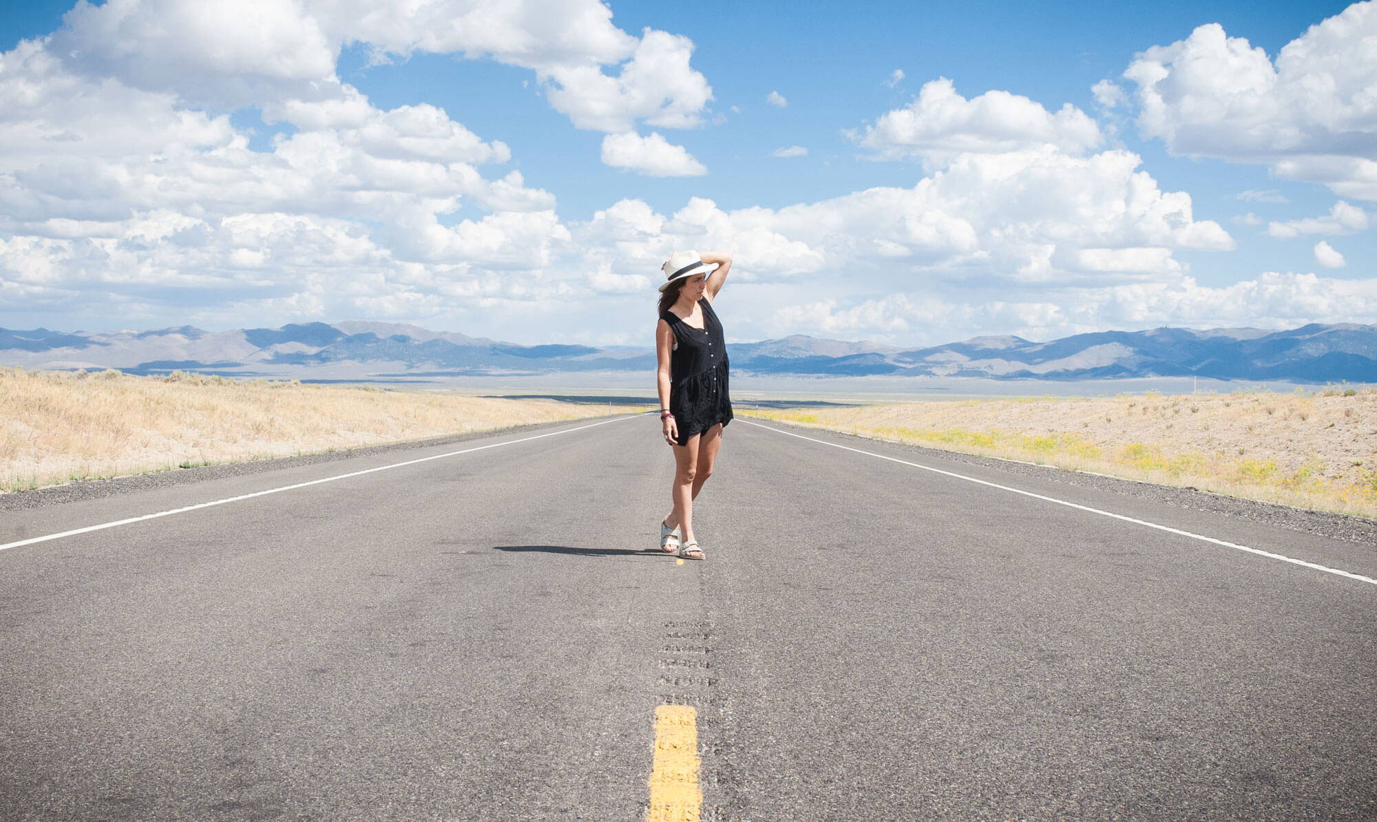 Woman wearing sun hat standing in the middle of the road with the sky and mountains behind her.