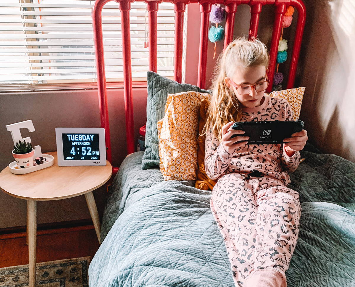 Emily Prior sits on bed playing device. A Dawn Clock sits on the bedside table.