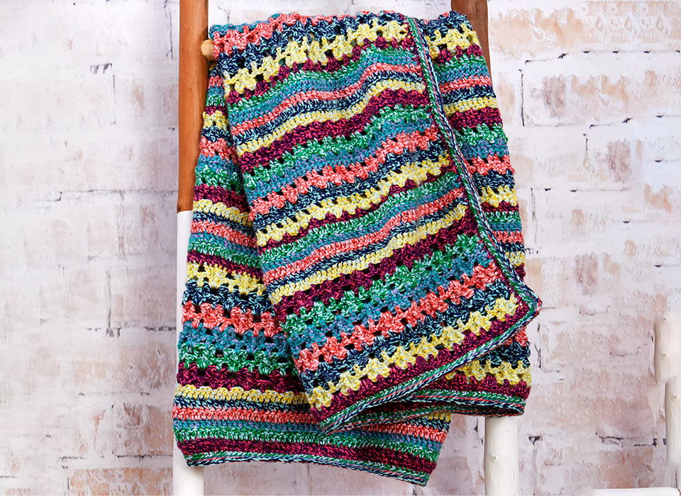 Colorful crocheted blanket draped over a wooden ladder against a textured background.