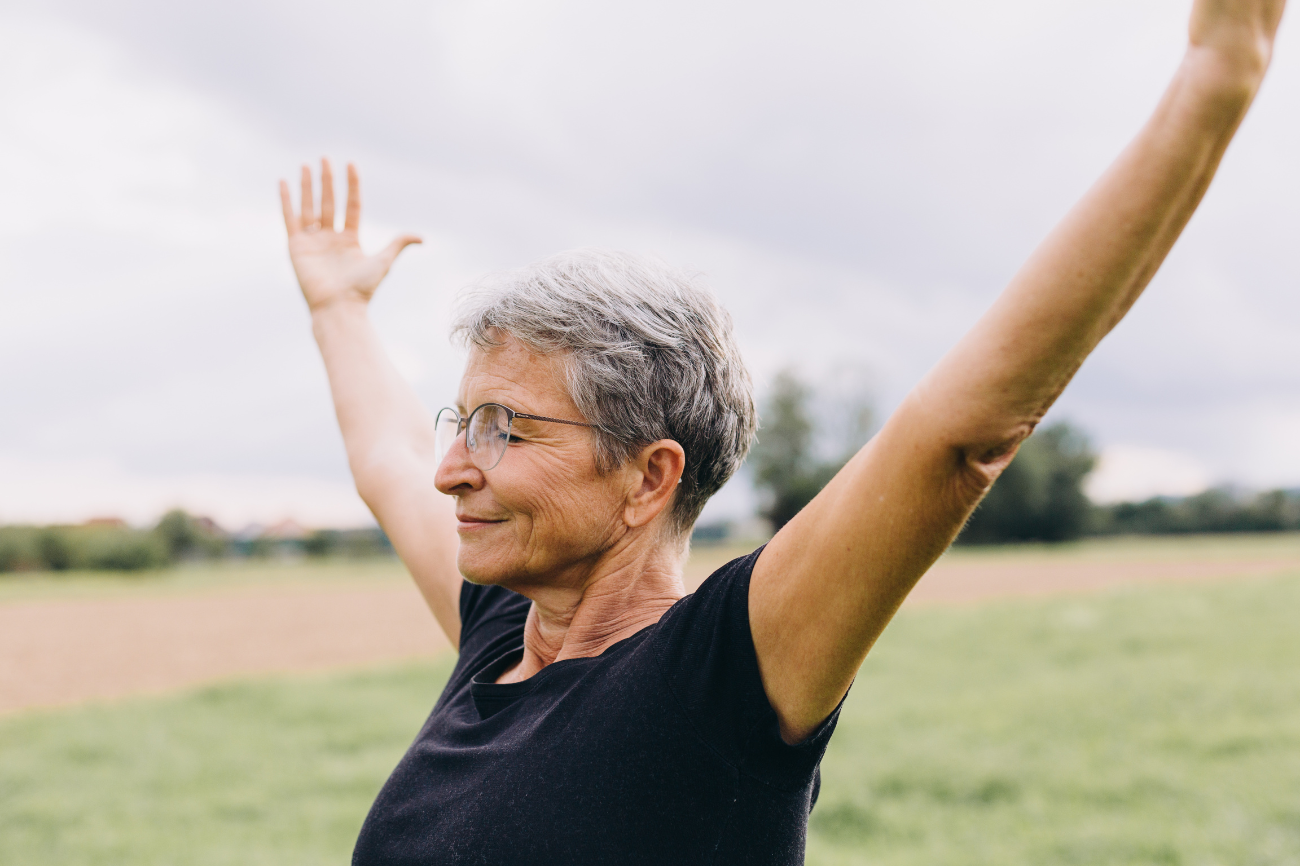 Older woman with short hair raising arms outdoors, smiling.