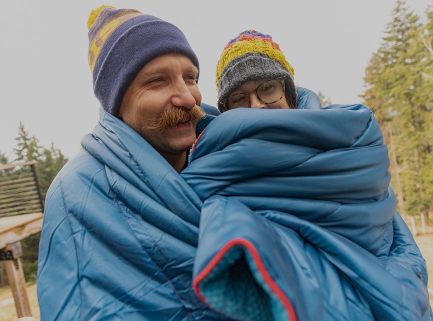 Two people bundled in blue sleeping bags, wearing colorful hats, outdoors.