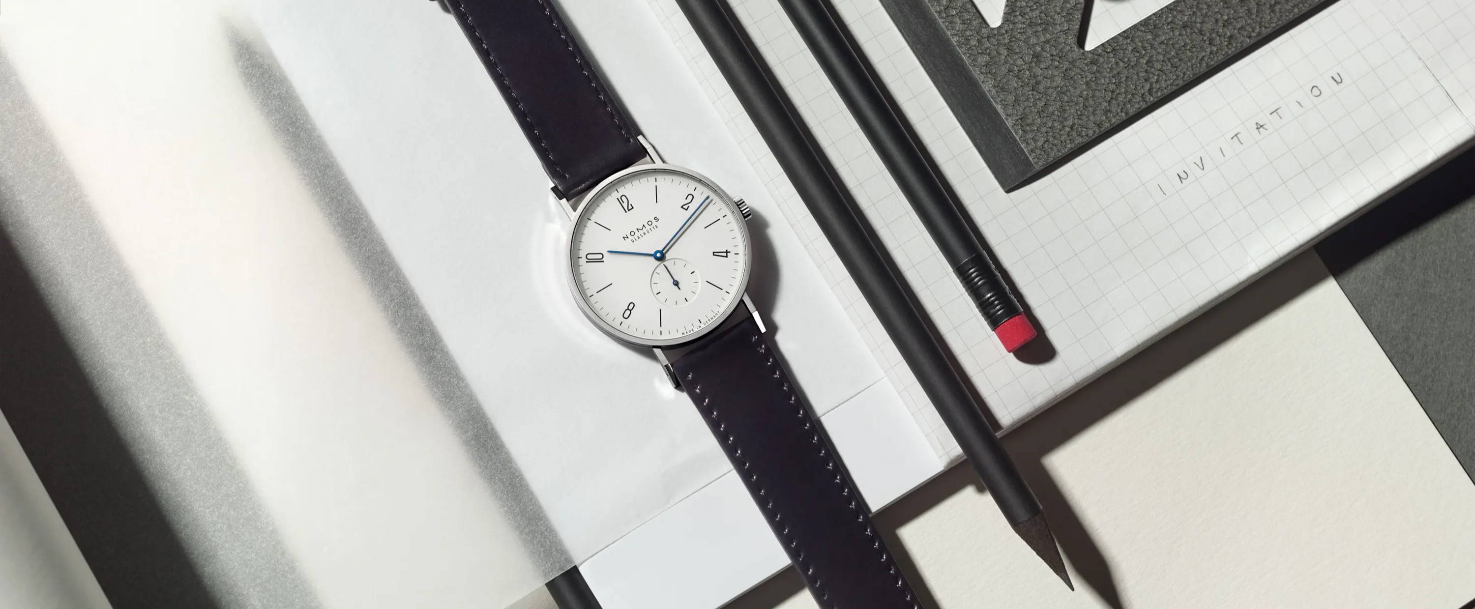 Nomos Glashutte Watch with Dark Leather Strap and White Dial on Notebook Next to Pencils