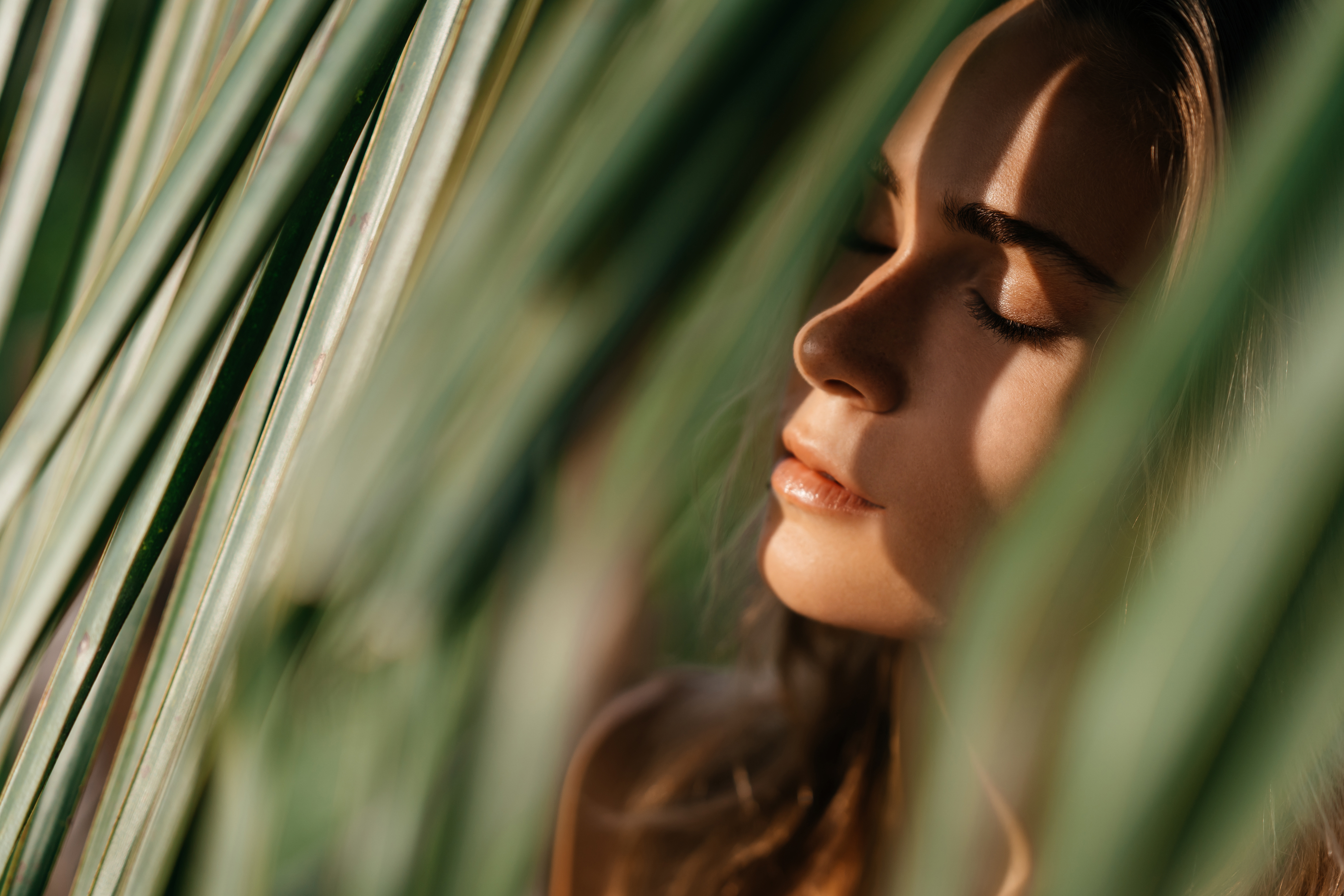 A woman with closed eyes surrounded by green leaves, softly lit.