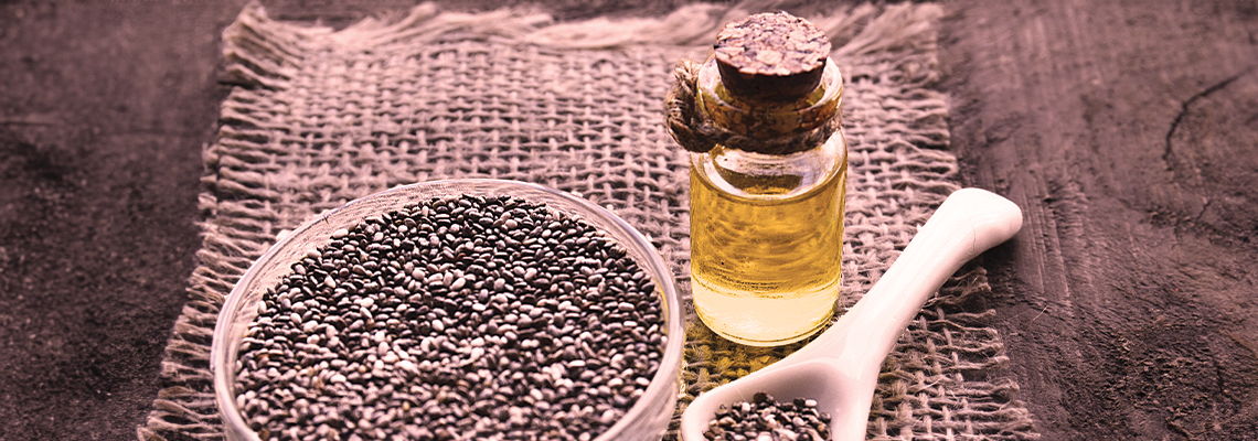 A bottle of oil beside a bowl of black seeds on a cloth.