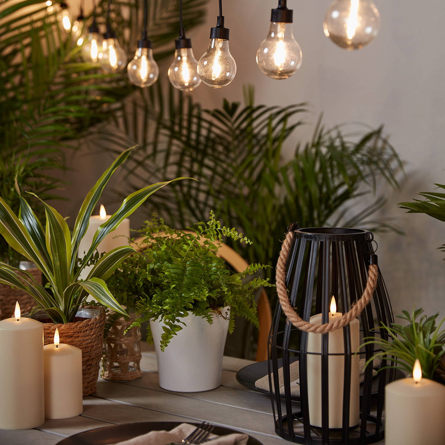 Une sélection de lanternes et bougies d'extérieur posée sur une table et une guirlande guinguette de style vintage suspendue en hauteur. 