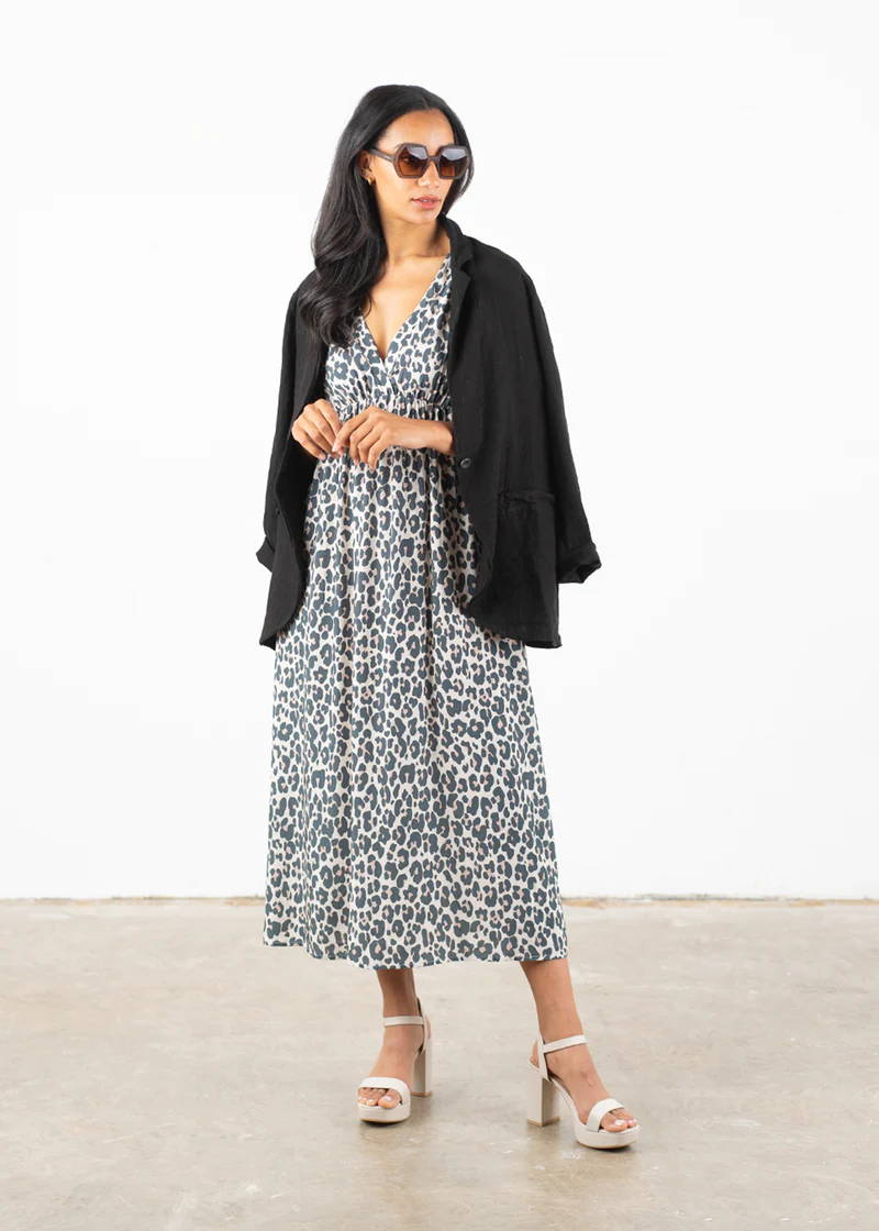 A model wearing a oatmeal midi dress with a leopard print pattern with a black linen jacket over her shoulders, black circular sunglasses and off white chunky heels