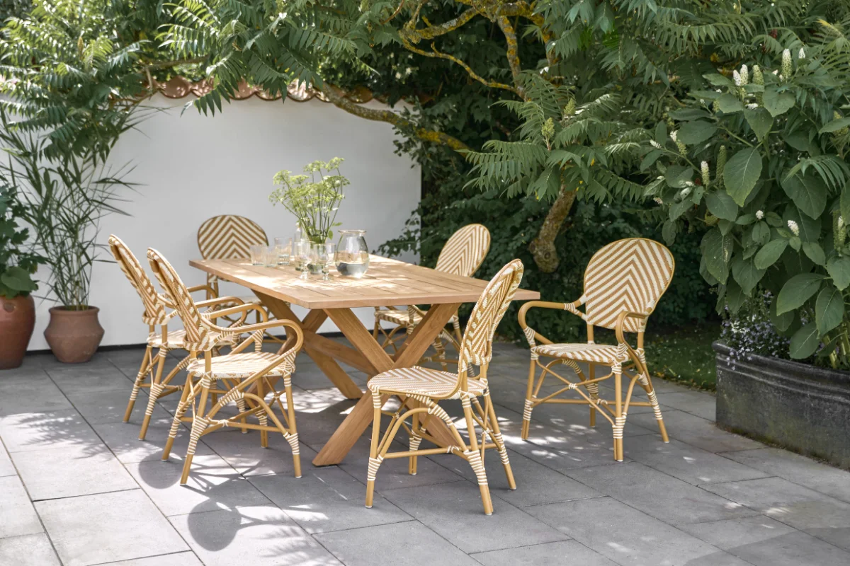 Outdoor dining set positioned beneath mature shade trees, creating a naturally cooled patio retreat with woven chairs and a teak dining table.