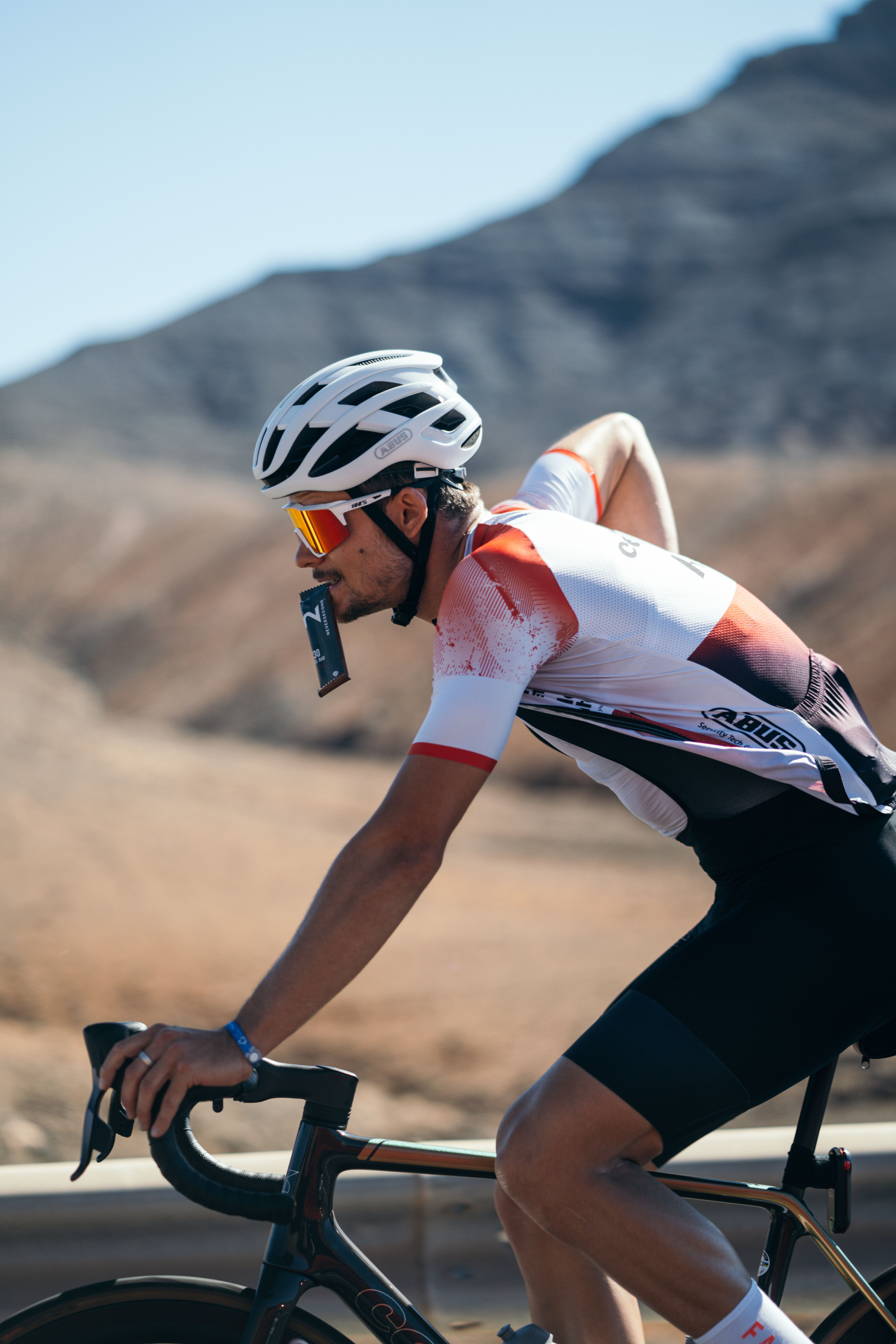 Triathlet Jan Stratmann auf dem Rennrad in bergiger Landschaft, mit einem Gel von Neversecond im Mund während der Fahrt.