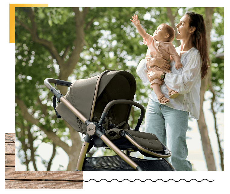 A woman holding a baby next to a stroller in a park.